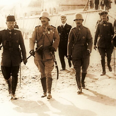 The image depicts a historical scene with a group of military personnel walking together in a line. They are dressed in formal military uniforms and accessories, including hats and medals, indicating a high rank or importance. The setting appears to be outdoors, possibly on a dirt or gravel path, with tents in the background suggesting a military camp. The overall tone of the photograph has a sepia or monochrome effect, conveying a sense of history. The individuals appear to be engaged in conversation or in a social setting, reflecting camaraderie among them.