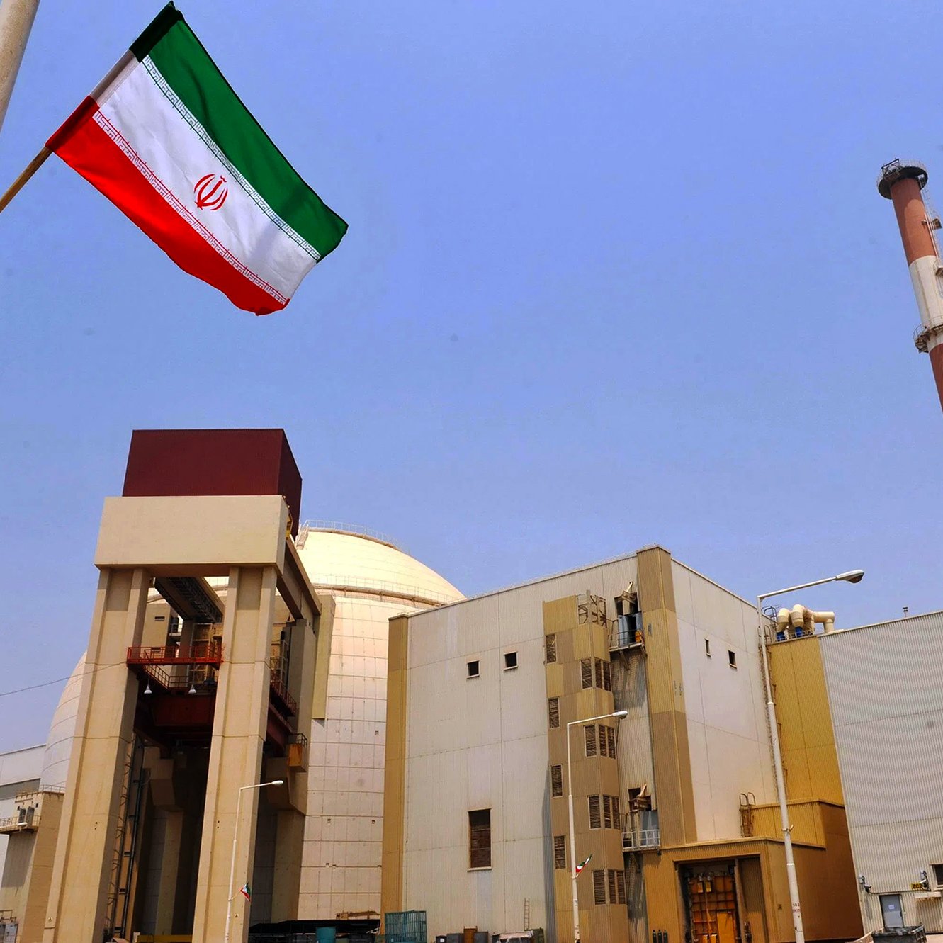L'image montre une installation industrielle avec des structures modernes. On peut voir une grande cheminée au fond et un bâtiment principal qui semble être lié à l'énergie. Au premier plan, il y a un mât avec le drapeau iranien flottant, qui arbore des bandes rouges, blanches et vertes. Le ciel est dégagé, créant un contraste avec les bâtiments. L'ensemble évoque une infrastructure liée à la production d'énergie, probablement nucléaire.