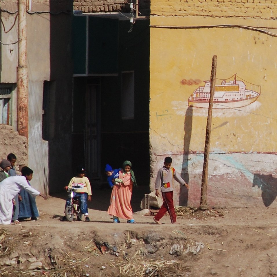 تظهر في الصورة مشهد لحياة يومية في منطقة شبه ريفية. في الجهة اليسرى يوجد شخص يرتدي ملابس بيضاء يحمل حقيبة، ويبدو أنه يسير نحو مجموعة من الأطفال والنساء. الأطفال يبدو أنهم يلعبون أو يتجهون إلى مكان ما. في الخلفية، توجد جدران ملونة تحتوي على رسوم جرافيتية وبعض الملابس المعلقة على حبل. الأرض تبدو جافة، مع بعض الحجارة والنباتات الصغيرة. يظهر كل ذلك الحياة البسيطة والمجتمع النشط في هذه المنطقة.