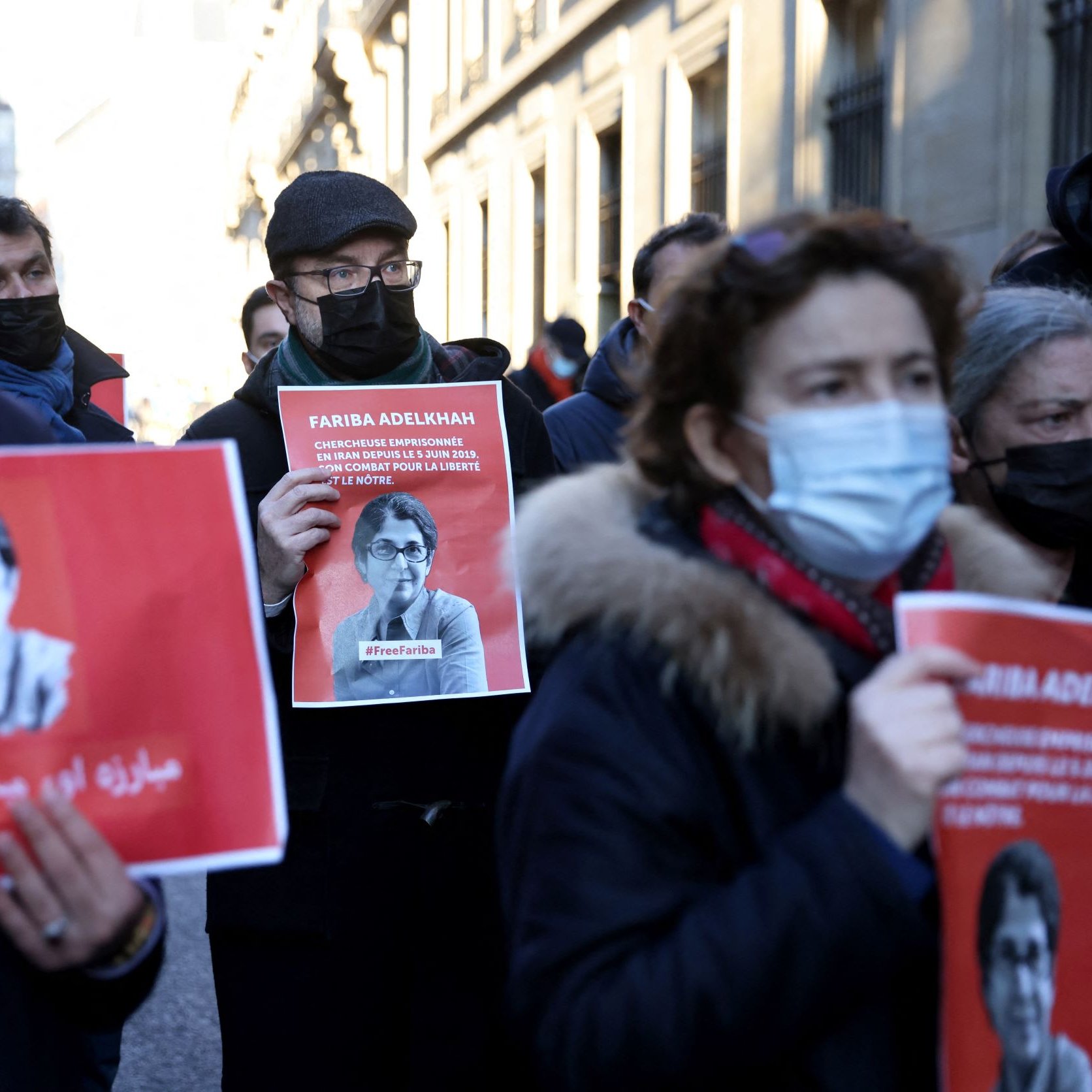 L'immagine mostra un gruppo di persone che partecipano a una manifestazione. Alcuni manifestanti tengono in mano cartelli con l'immagine e il nome di una persona, accompagnati da slogan scritti in arabo e in italiano. La maggior parte delle persone indossa mascherine e abbigliamento invernale, suggerendo che l'evento si svolge in un periodo freddo. L'atmosfera sembra essere seria e determinata, con un focus sulla questione rappresentata dai cartelli.