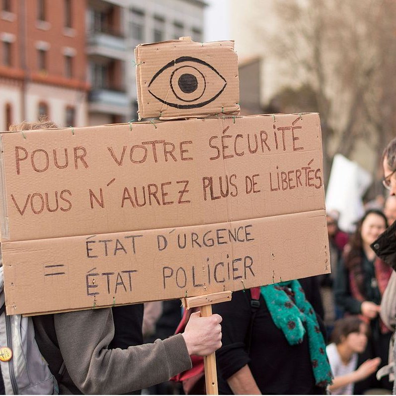 L'image représente une manifestation où une personne tient une pancarte en carton. Sur cette pancarte, on peut lire un message dénonçant la restriction des libertés au nom de la sécurité. Le symbole d'un œil est visible en haut de la pancarte, renforçant le thème de la surveillance. Les participants de la manifestation semblent mobilisés, et il y a une ambiance de protestation dans l'air.