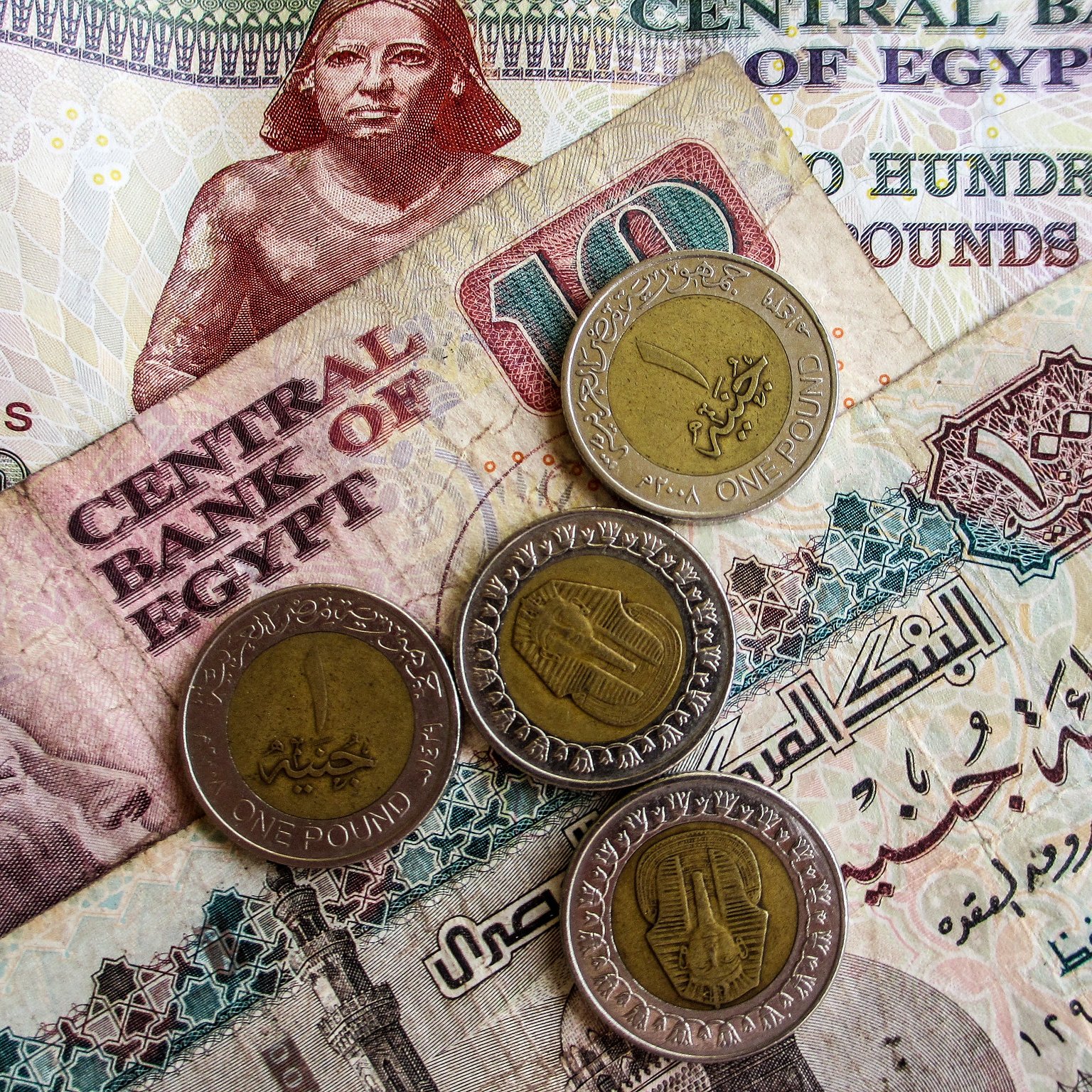 تظهر في الصورة مجموعة من العملات المصرية. هناك ورقة نقدية من فئة المائة جنيه بالإضافة إلى فئة العشرة جنيه، مع عدد من العملات المعدنية من فئة الجنيه الواحد. التصميمات المختلفة توضح رموز ومعالم ثقافية مرتبطة بمصر، مما يعكس التنوع في العملة المصرية.