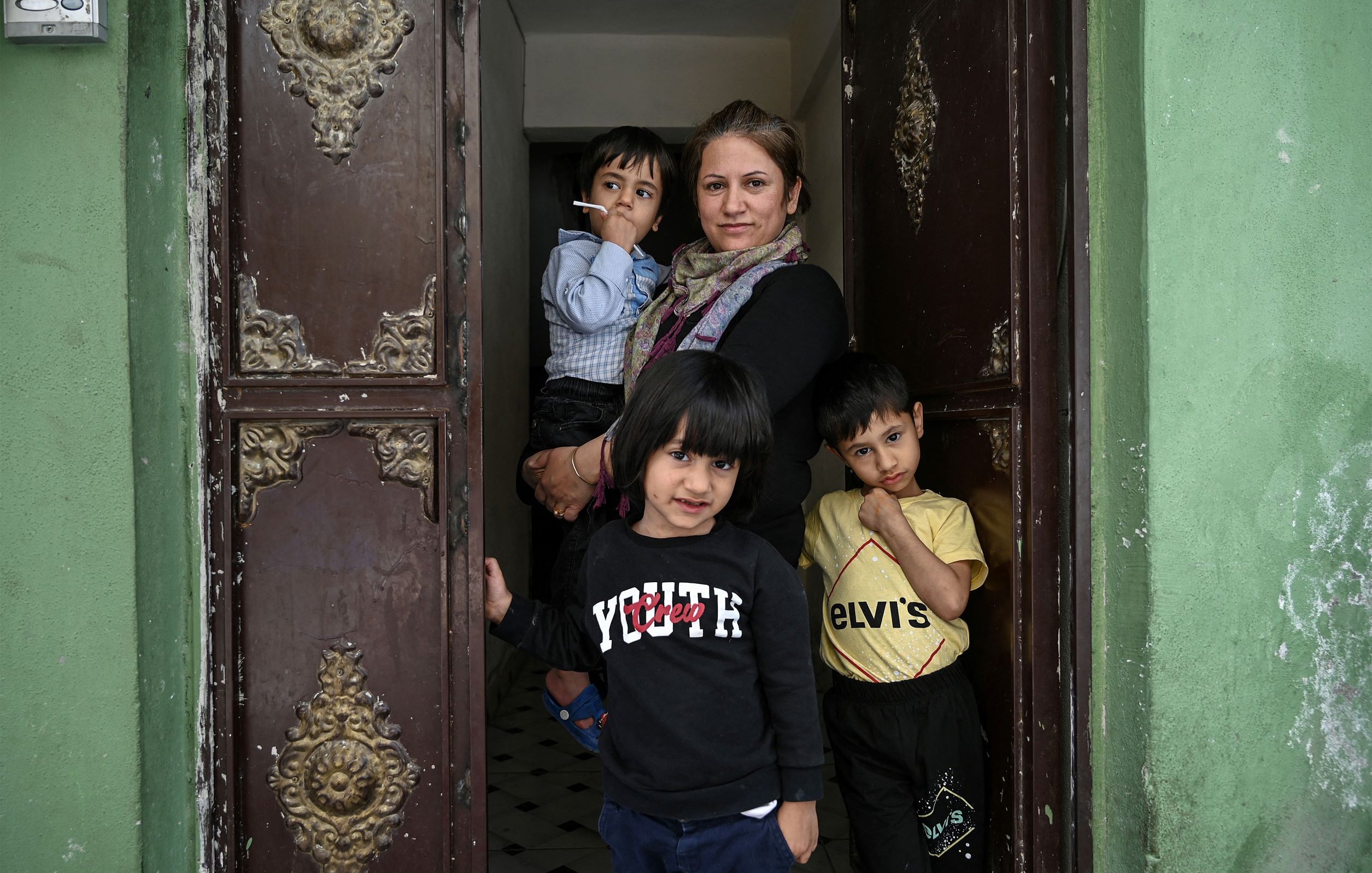 L'image montre une femme debout dans une porte d'entrée, entourée de trois jeunes enfants. Elle a un air calme et tient une cigarette. Les enfants semblent curieux et sont vêtus de vêtements décontractés. Leurs expressions varient, certains souriant tandis que d'autres regardent vers l'objectif. Le fond présente des murs verts et une porte ornée de détails décoratifs, ajoutant une touche particulière à la scène.