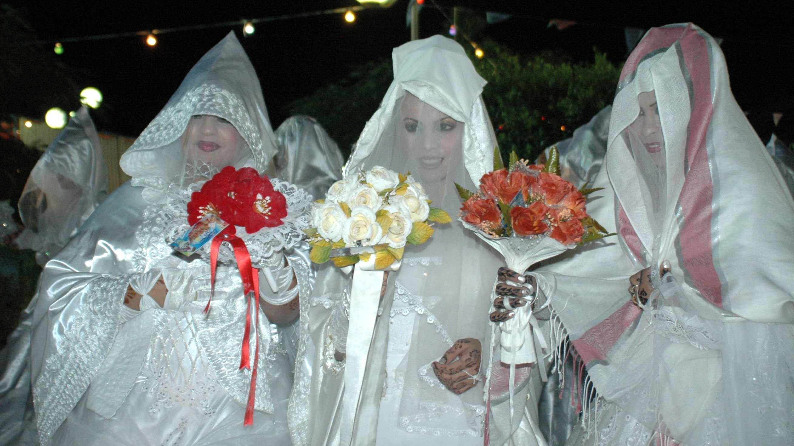 The image features three women dressed in elaborate white outfits that resemble bridal attire. They are wearing head coverings and are holding bouquets of flowers. The setting appears to be outdoors at night, illuminated by soft lights above them. The overall atmosphere seems festive, possibly related to a celebration or cultural event. The women's faces are mostly obscured, adding a mysterious or dramatic effect to the scene.