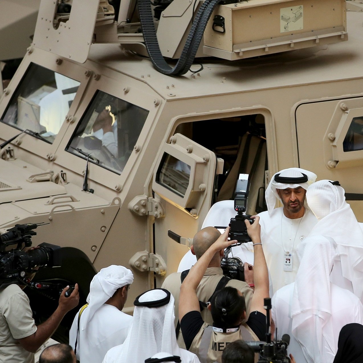 در این تصویر، گروهی از مردان به لباس سنتی عربی، در حال برگزاری یک رویداد یا نمایشگاه هستند. یکی از افراد در مرکز تصویر به نظر می‌رسد که در حال صحبت با دیگران است و چند نفر دیگر نیز در حال عکس‌برداری هستند. در پس‌زمینه، یک وسیله نقلیه نظامی با رنگ شن و طراحی خاص دیده می‌شود. این صحنه نشان‌دهنده یک موقعیت رسمی و احتمالا نظامی است، با حضور رسانه‌ها و توجه به جزئیات.