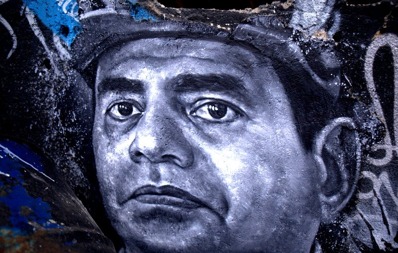 تظهر الصورة رسما جداريًا لوجه رجل، حيث يتميز التعبير بالعمق والتفاصيل الدقيقة. يُظهر الرسم عيونًا تعكس مشاعر قوية، والخطوط تتباين بين الظل والنور، مما يعطي إحساسًا بالحياة والواقعية. الخلفية تحتوي على ألوان داكنة ومخططات فنية أخرى، مما يبرز التركيز على وجه الرجل ويعزز من جمال العمل الفني.