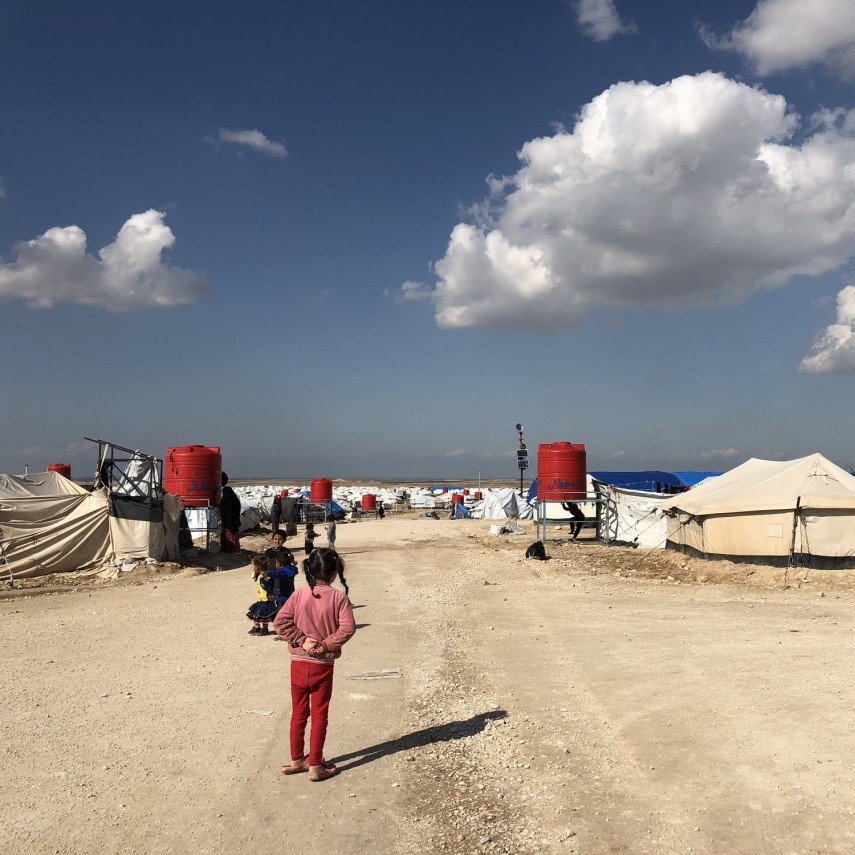 L'image montre un camp de réfugiés avec des tentes disposées de part et d'autre d'un chemin de terre. Au fond, on aperçoit des nuages blancs dans un ciel bleu. Des enfants sont présents, certains semblent jouer ou se déplacer dans le camp. Les abris sont principalement en toile, avec quelques réservoirs d'eau visibles. L'atmosphère paraît calme malgré les conditions de vie apparentes de ces lieux.