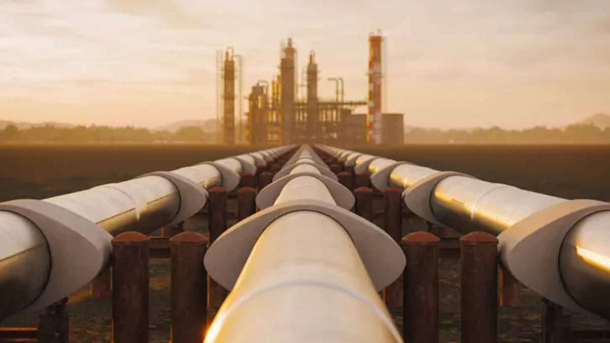 The image features a series of large pipelines leading towards a distant industrial facility, likely a refinery or processing plant. The foreground showcases the pipes arranged in a linear perspective, drawing the viewer's eye toward the structure in the background. The setting is illuminated by a warm, golden light, suggesting either sunrise or sunset, which creates an atmospheric and industrial ambiance. The landscape around the pipes appears open, possibly indicating a rural or undeveloped area.