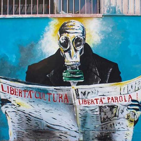 L'immagine rappresenta un murale che ritrae una figura umana con una maschera antigas, seduta e intenta a leggere un quotidiano. Il soggetto indossa un abbigliamento scuro e la scena è caratterizzata da una palette di colori vivaci, con sfumature di blu e bianco sullo sfondo. Le parole "LIBERTÀ", "CULTURA" e "LIBERTÀ di PAROLA!" sono scritte con un carattere forte e chiaro, sottolineando un messaggio di libertà e critica sociale. La data "2014" indica quando è stata realizzata l'opera, con il riferimento a "Cuba Libera" che suggerisce un legame con temi di libertà e diritti civili.