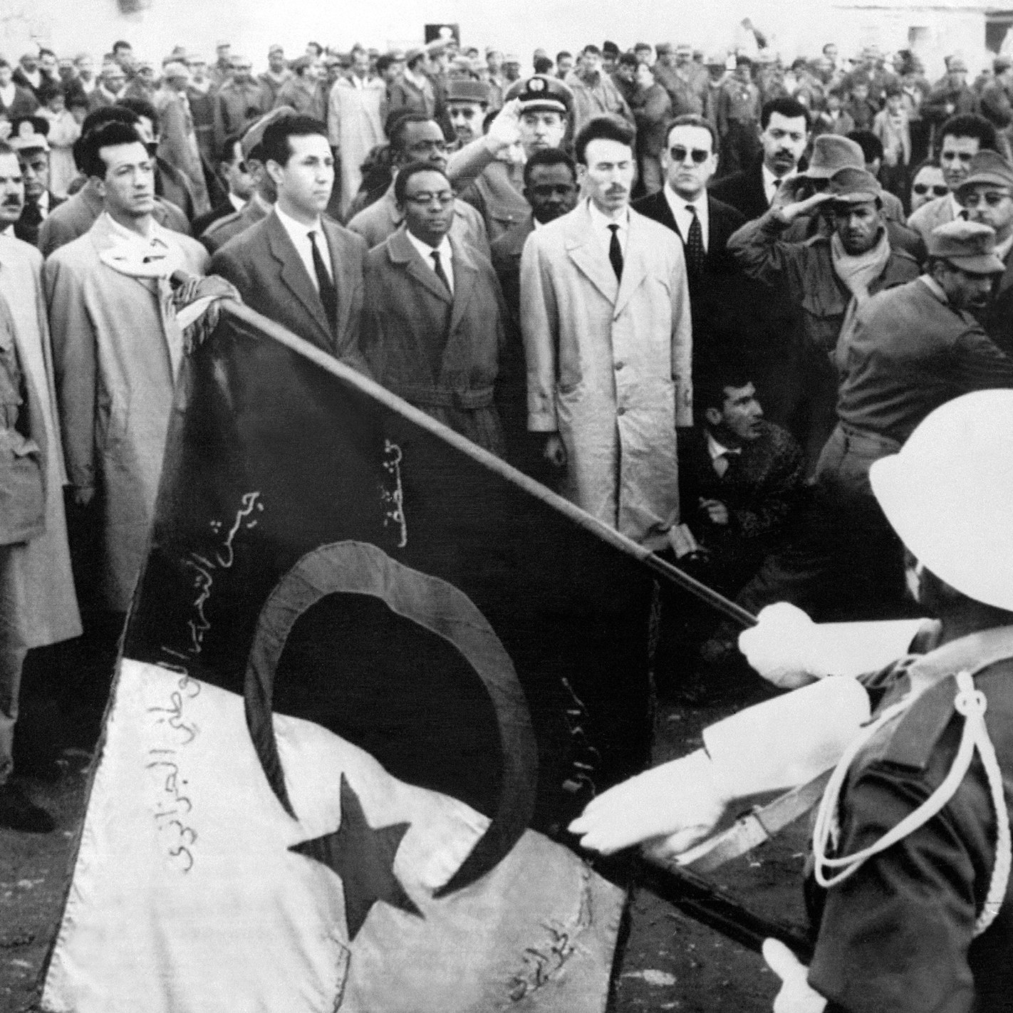 The image depicts a historical scene with a large crowd gathered, likely for a ceremonial or significant event. In the foreground, two soldiers are saluting while holding a flag, which features a crescent and a star, elements often associated with some national flags. The crowd appears to consist of men in various types of clothing, with some wearing military uniforms and others in suits. The overall atmosphere suggests a moment of national importance, possibly related to independence or a political event.