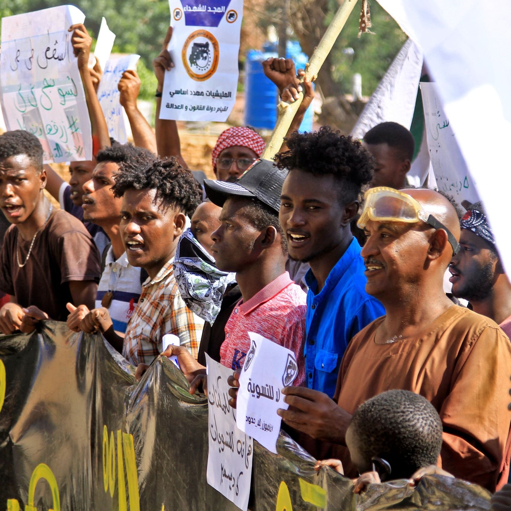 L'image montre un groupe de manifestants rassemblés, brandissant des pancartes et exprimant des slogans. Ils semblent engagés et passionnés, avec des expressions faciales intenses. Les gens portent des vêtements variés et il y a des drapeaux ou des bannières en arrière-plan. L'ambiance générale semble être celle d'une manifestation pour une cause précise.