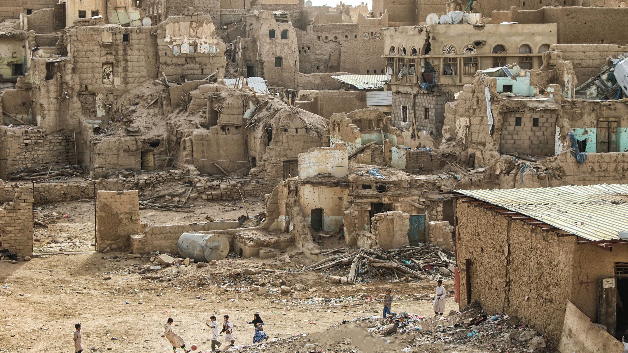 L'image montre un paysage urbain en ruines, typique d'une zone désertique. On peut voir des bâtiments en terre et en briques, avec des murs partiellement effondrés. Les structures semblent abandonnées et en mauvais état, certaines présentant des ouvertures et des décombres. Au premier plan, plusieurs enfants jouent, ce qui apporte une touche de vie à ce décor désertique. Le sol est jonché de déchets, et le ciel est clair, ce qui accentue l'aspect aride de l'environnement.