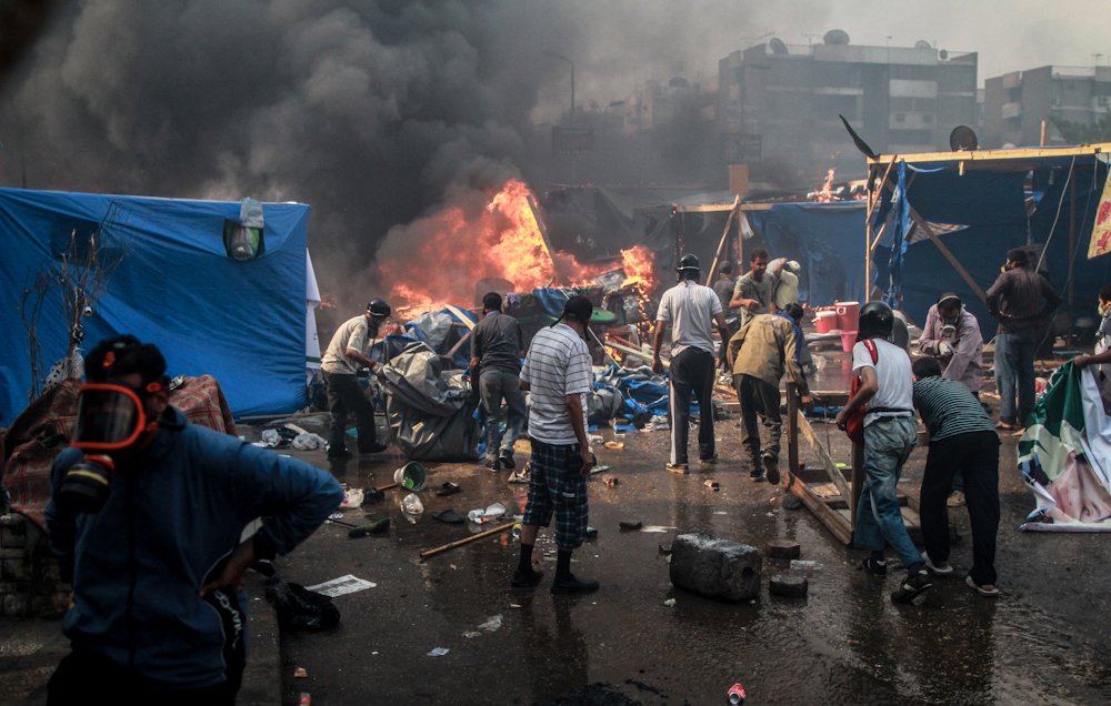 L'image montre une scène de chaos et de destruction. On peut voir des personnes rassemblées autour de flammes, visiblement en train de fuir ou de lutter contre un incendie. La fumée noire s'élève dans le ciel, obscurcissant la vue. Des tentes et des objets sont dispersés dans l'environnement, suggérant une occupation ou une manifestation. La situation semble tendue, avec un sentiment d'urgence parmi les personnes présentes.