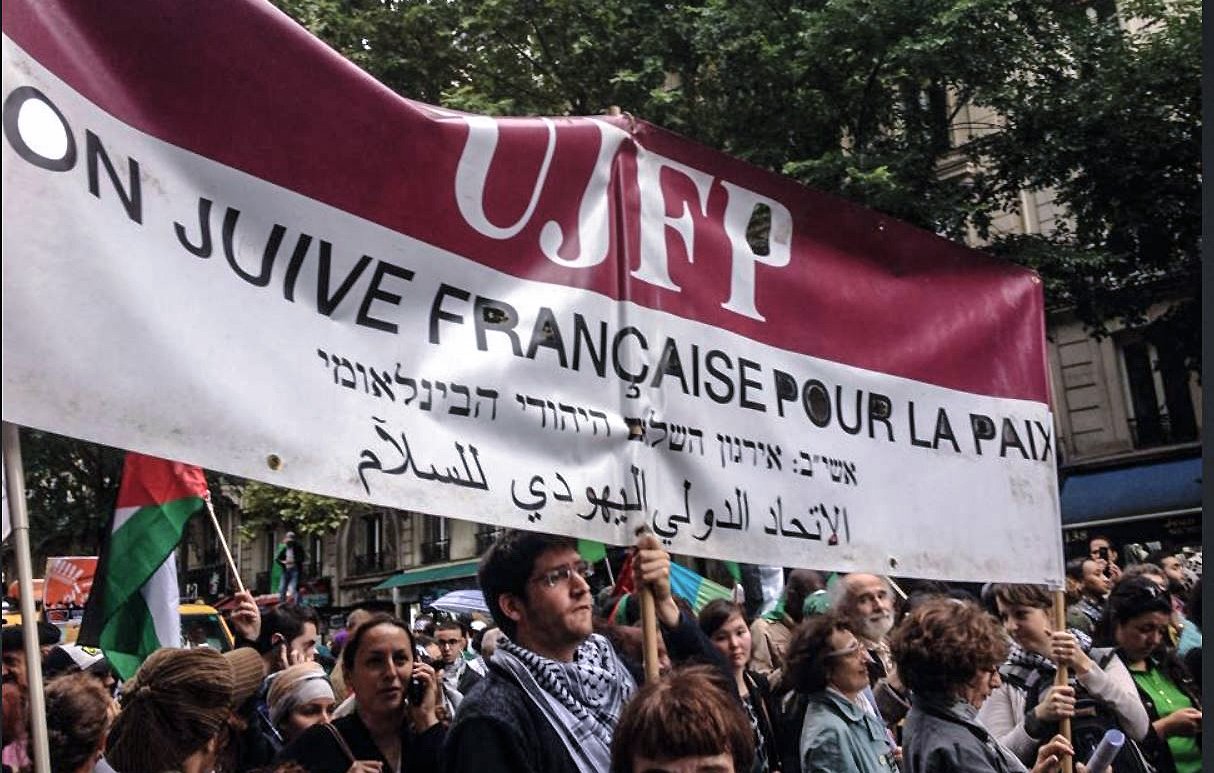 L'image montre une manifestation où des personnes tiennent une grande banderole. Cette banderole mentionne "UJFP" et affiche des messages en français, hébreu et arabe, qui semblent appeler à la paix. Le contexte suggère un rassemblement pour des causes sociales ou politiques, probablement en lien avec la paix au Moyen-Orient. En arrière-plan, on voit une foule de manifestants, des arbres et des bâtiments, ce qui indique que l'événement se déroule en milieu urbain.