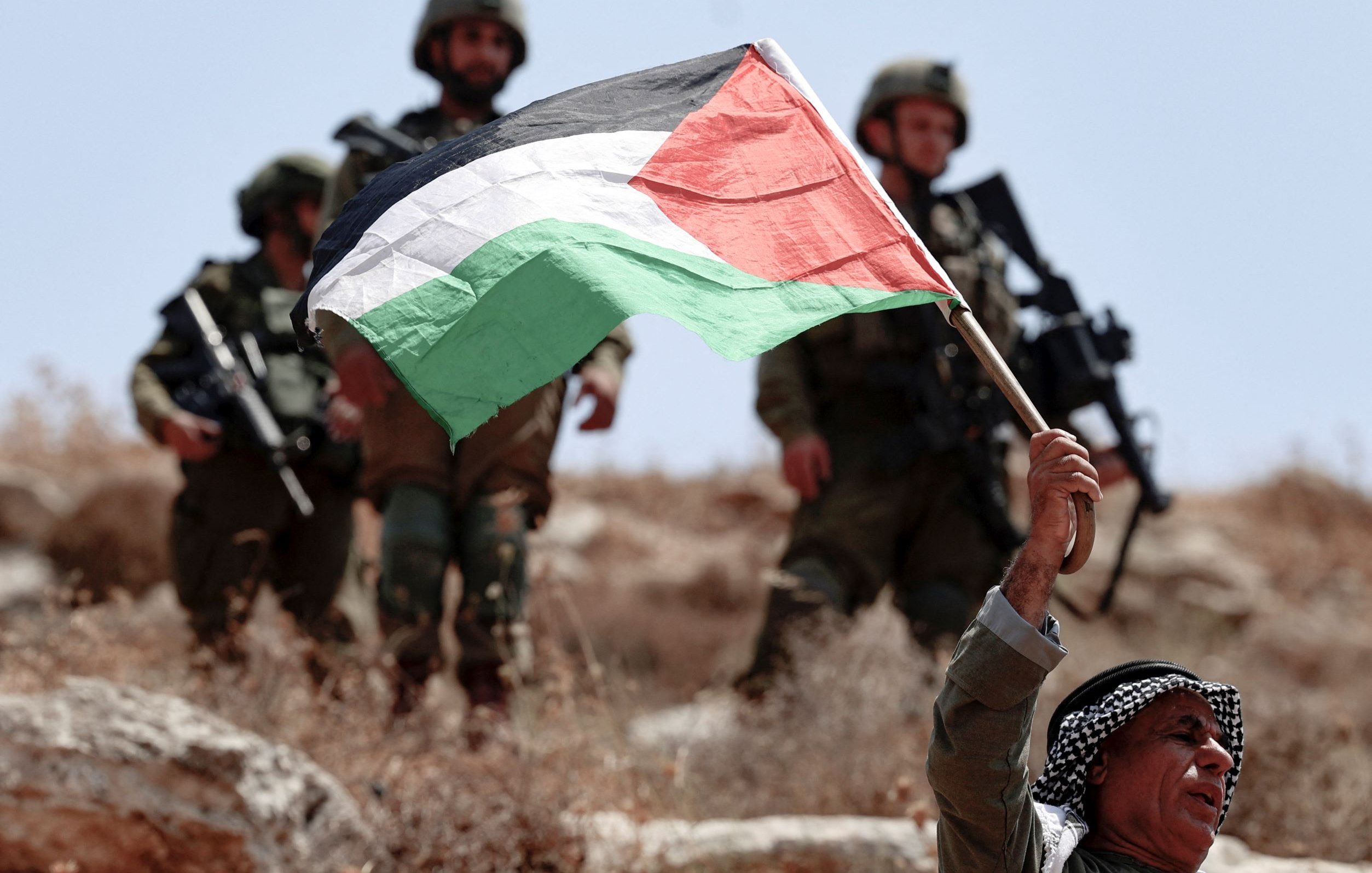 L'image montre un homme brandissant fièrement un drapeau palestinien. Il semble être en première ligne, tandis que des soldats en arrière-plan, probablement des soldats israéliens, sont visibles. Le paysage est sec et rocailleux, suggérant un environnement rural ou semi-aride. L'atmosphère semble tendue en raison de la présence militaire et du geste de protestation de l'homme.