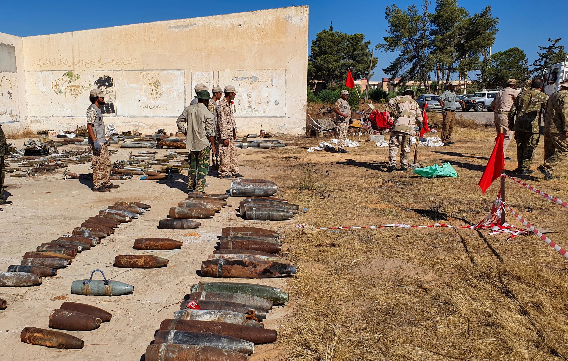 تظهر الصورة مجموعة من الأشخاص يرتدون زيًا عسكريًا، يتواجدون في منطقة مفتوحة تحتوي على العديد من القذائف والذخائر القديمة المصفوفة على الأرض. الخلفية تحتوي على جدران مبنى متهدم، بينما تظهر شجيرات وأشجار في المنطقة المحيطة. يبدو أن هناك بعض الأعلام والأعلام الحمراء في الموقع، مما يشير إلى نشاطات أو إجراءات تتعلق بالسلامة أو التفكيك. الجو العام يوحي بأن هناك عملية تُجرى للتعامل مع هذه الذخائر.