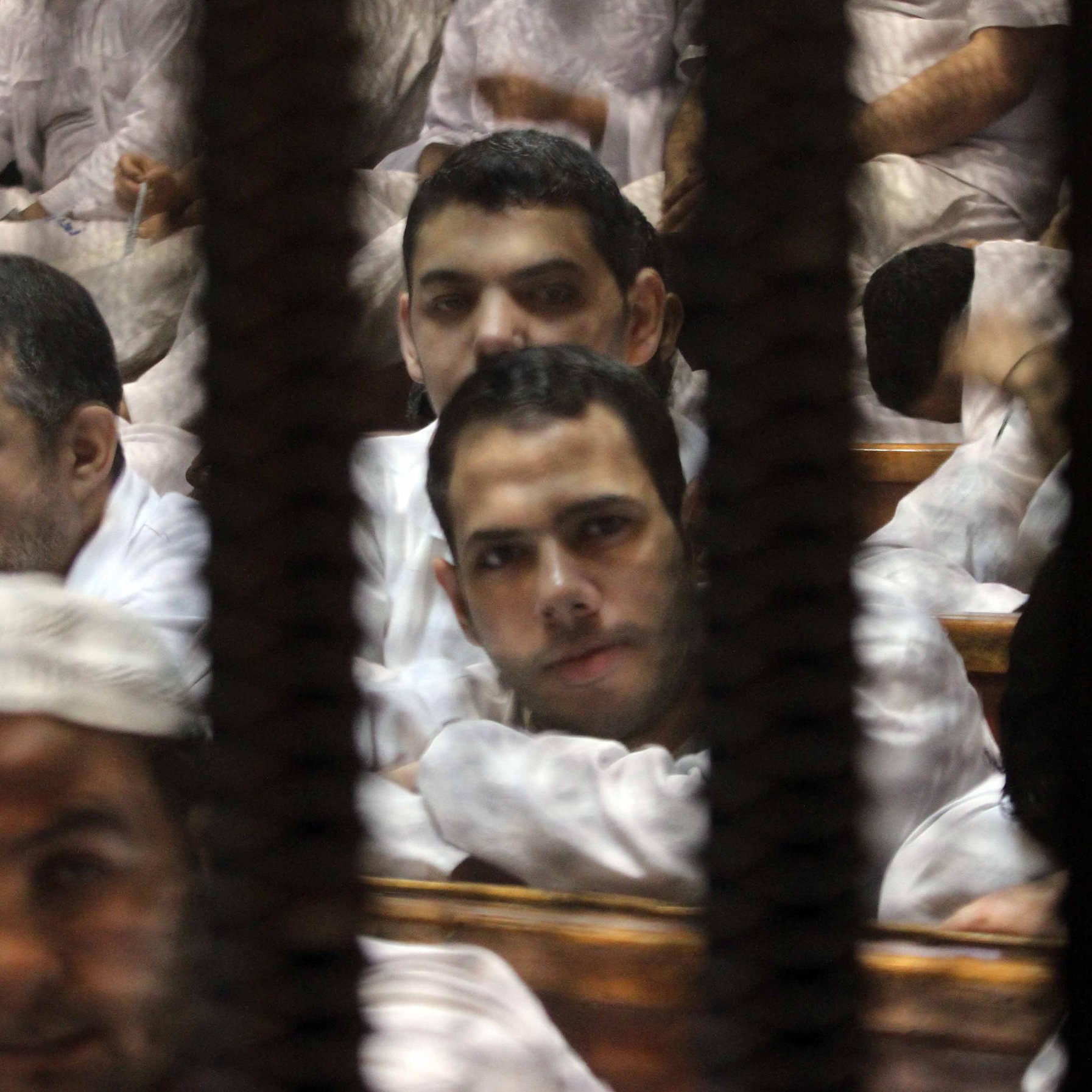 L'image montre un groupe d'hommes assis dans une salle, probablement lors d'une audience judiciaire. Ils portent des vêtements simples, généralement de couleur claire. Leurs expressions semblent sérieuses, et ils sont entourés de barreaux, ce qui suggère un environnement de détention ou de tribunal. L'ambiance générale est tendue, et la composition de la photo cadre bien les visages des hommes à travers les barreaux.