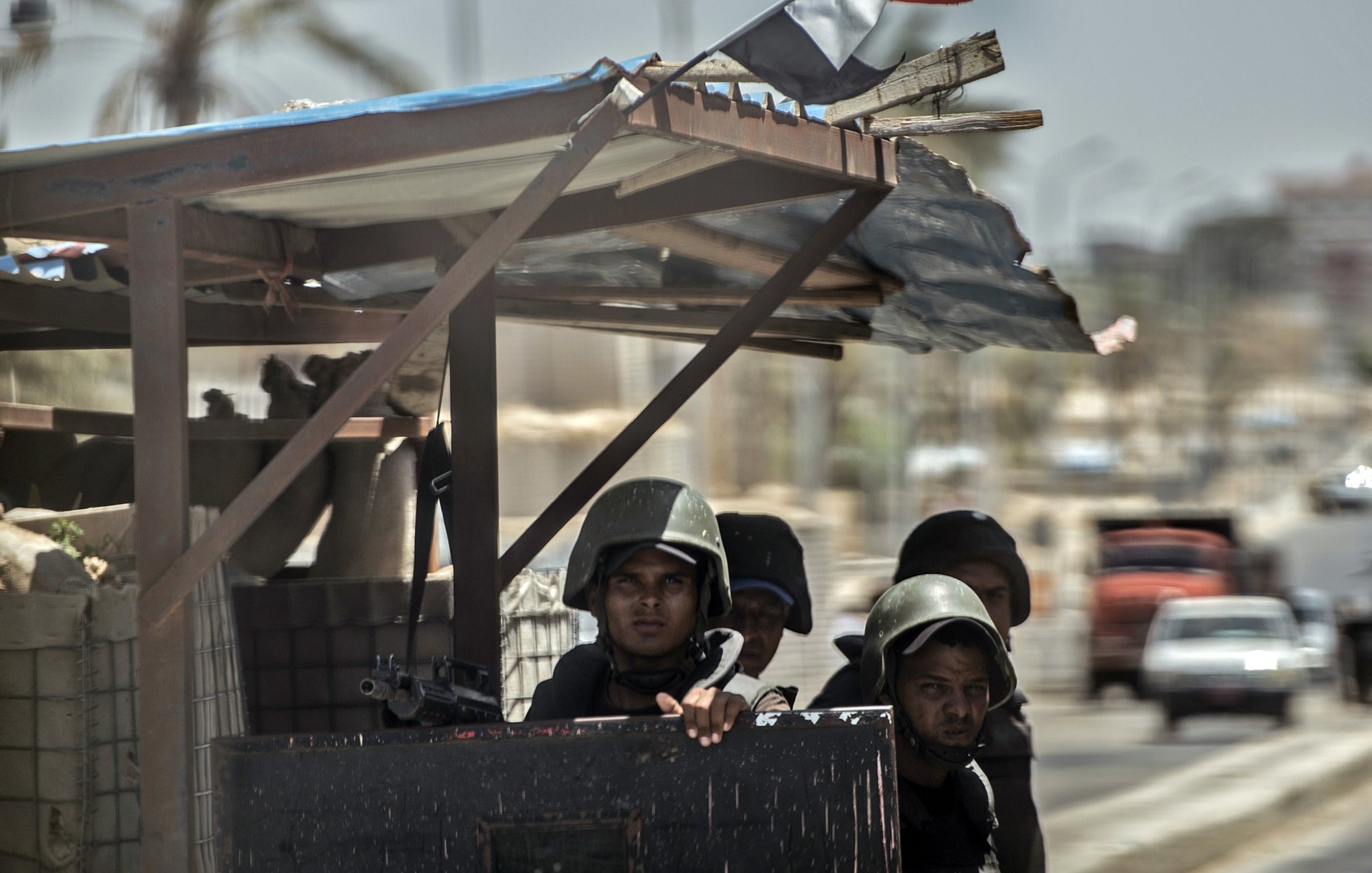 The image depicts a group of soldiers stationed at a military outpost. They are wearing helmets and carrying weapons, suggesting they are on alert or guarding a specific area. The backdrop includes palm trees and some vehicles, indicating an urban environment. A flag can be seen on top of the structure, adding to the military context of the scene. The overall atmosphere appears tense and focused on security.