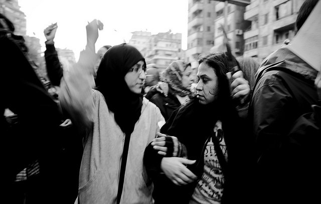 L'image montre un groupe de personnes lors d'une manifestation. Deux jeunes femmes, l'une portant un hijab et l'autre les cheveux détachés, semblent échanger des mots tout en levant les bras, ce qui indique un sentiment d'engagement et de solidarité. L'ambiance est dynamique, et le noir et blanc donne une atmosphère intense à la scène. En arrière-plan, on peut apercevoir d'autres manifestants, créant une impression de mouvement et de rassemblement.