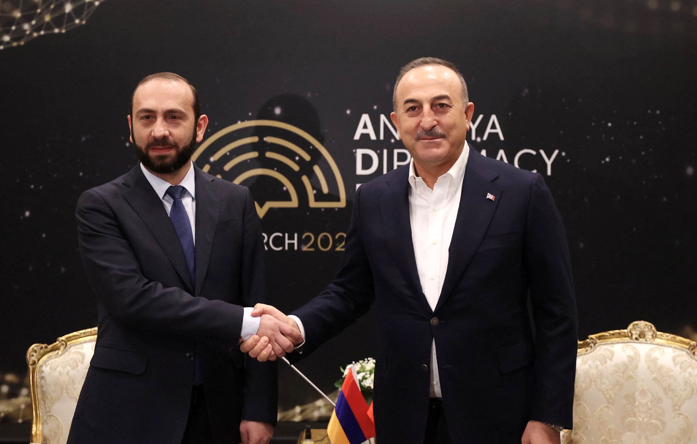 تظهر الصورة شخصين يتصافحان في مناسبة دبلوماسية. يقف الشخصان أمام خلفية تحمل عبارة "أنقرة دبلوماسية" وتاريخ مارس 2022. يرتدي أحدهما بدلة رسمية بينما يبدو الآخر أيضًا في زي رسمي. المكان يبدو أنيقًا، ويوجد خلفهما تصميم مميز يبرز الطابع الرسمي للاجتماع.