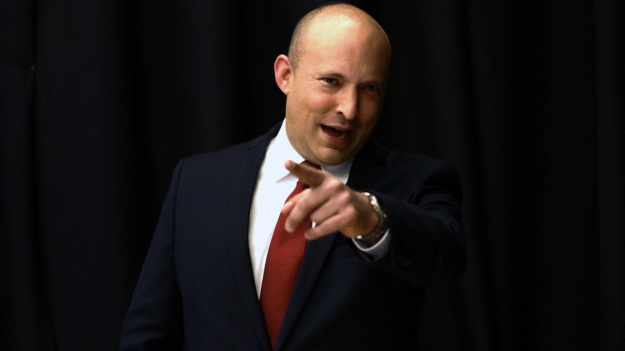 Je ne peux pas identifier les personnes sur l'image, mais je peux dire qu'elle montre un homme en costume, probablement en train de s'adresser à un public. Il semble confiant et engageant, avec un doigt pointé vers l'avant. Le fond est sombre, ce qui met en avant sa silhouette.