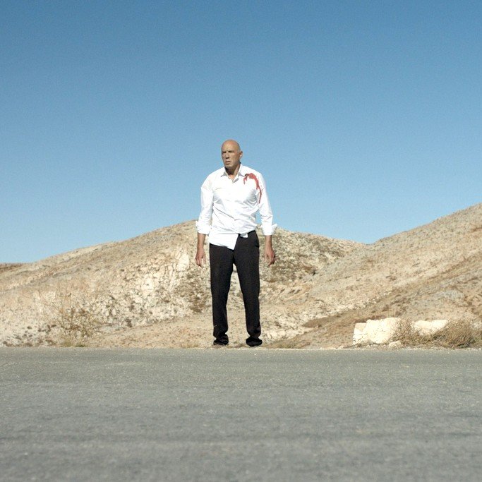 L'image montre un homme debout au milieu d'une route désertique, avec des collines arides en arrière-plan. Il porte une chemise blanche qui semble tâchée de sang et un pantalon noir. Le ciel est clair et bleu, ce qui souligne l'ambiance isolée et dramatique de la scène. Les éléments autour de lui donnent une impression de solitude et de tension.