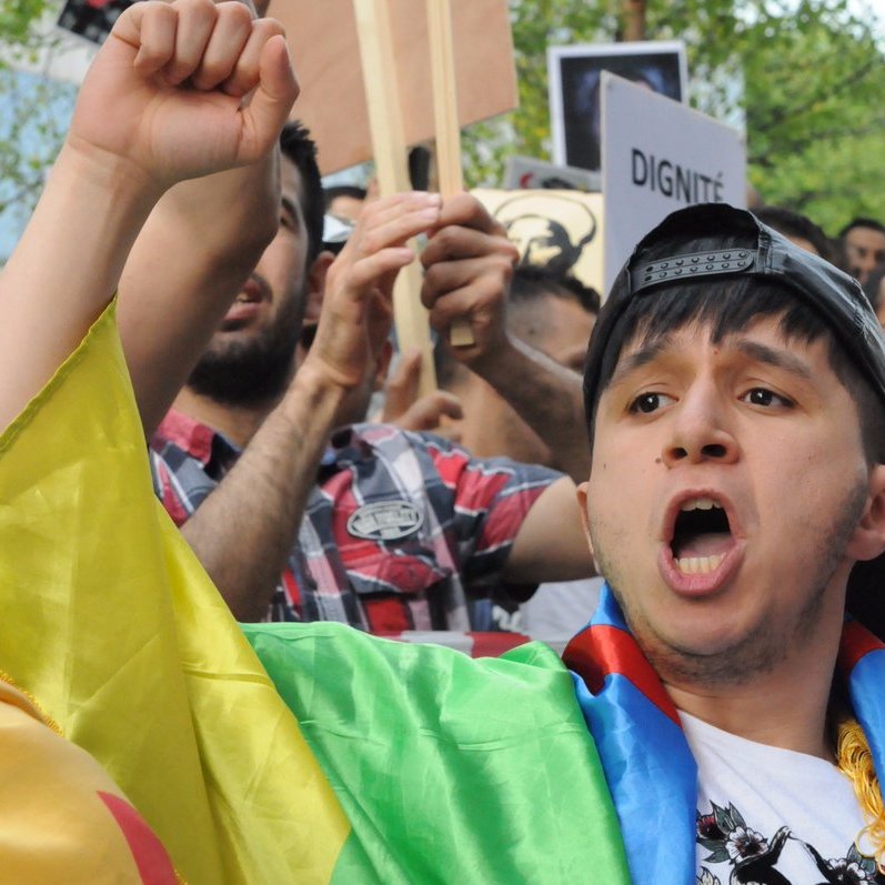 L'image montre une scène de manifestation, où un jeune homme, portant un drapeau coloré, crie avec passion. Il semble mobilisé et engagé dans la cause pour laquelle les manifestants se sont rassemblés. En arrière-plan, on peut voir d'autres personnes tenant des pancartes et exprimant leurs préoccupations. L'atmosphère est remplie d'énergie et de détermination.