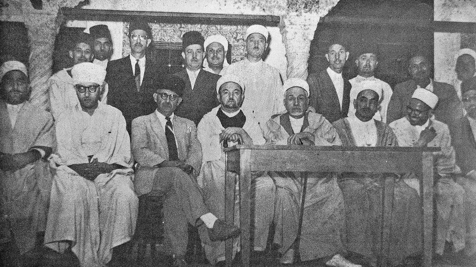 L'image montre un groupe de personnes rassemblées dans une pièce. Elles portent des vêtements traditionnels, notamment des turbans et des robes longues. Au centre, quelques individus sont assis à une table, tandis que d'autres se tiennent debout derrière eux. L'atmosphère semble formelle et solennelle, suggérant une réunion ou une cérémonie. Les murs sont en pierre, ce qui donne un aspect historique à l'ensemble.