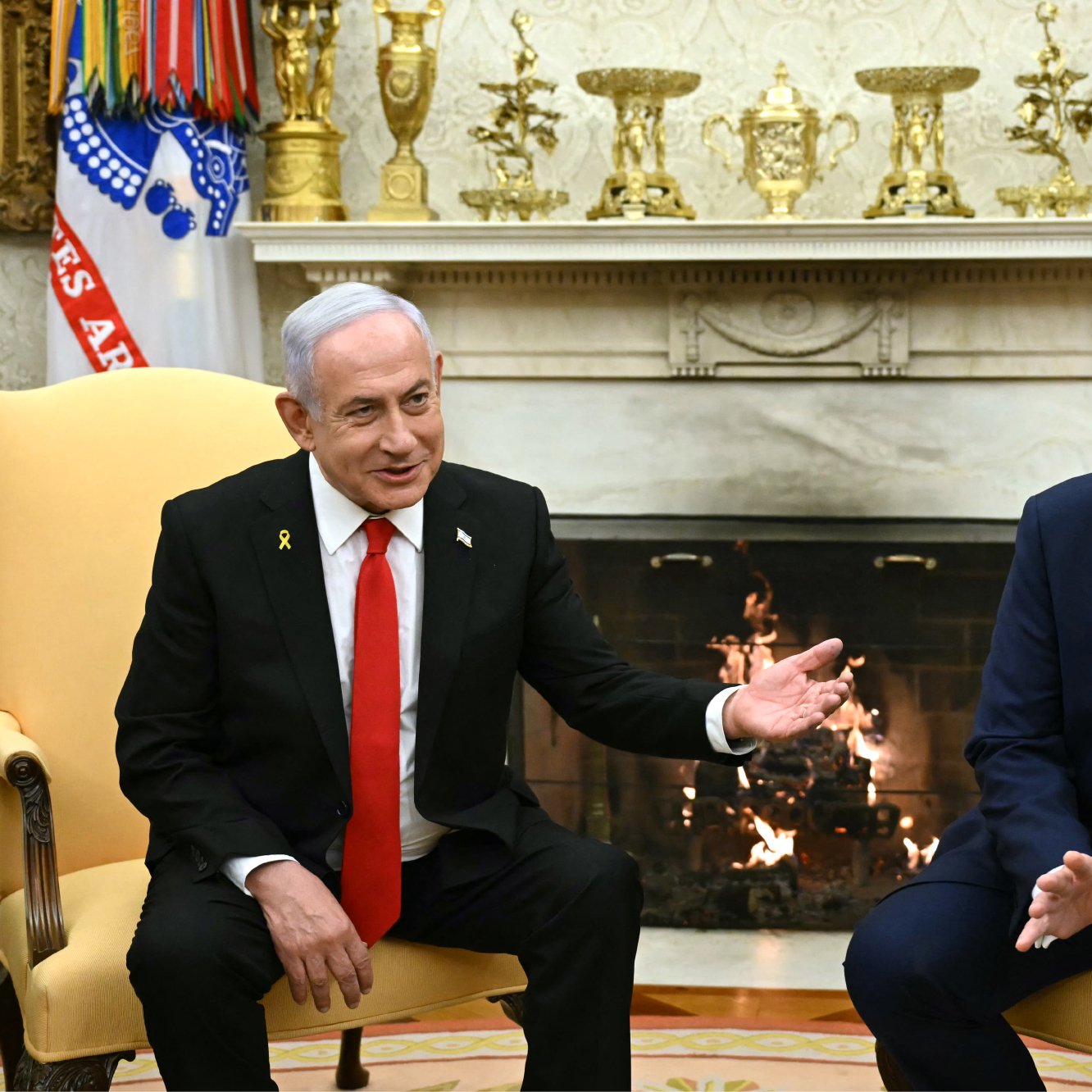 در این تصویر، دو شخصیت سیاسی در یک اتاق ملاقات نشسته‌اند. یکی از آنها در لباس رسمی سیاه و با یقه سفید و کراوات قرمز حاضر شده است و دیگری در کت و شلوار تیره و با کراوات آبی نشسته است. پس‌زمینه شامل یک شومینه و دکوراسیون کلاسیک است. هر دو شخصیت در حال گفت‌وگو به نظر می‌رسند و حرکات دست آنان نشان‌دهنده تبادل نظرات است.