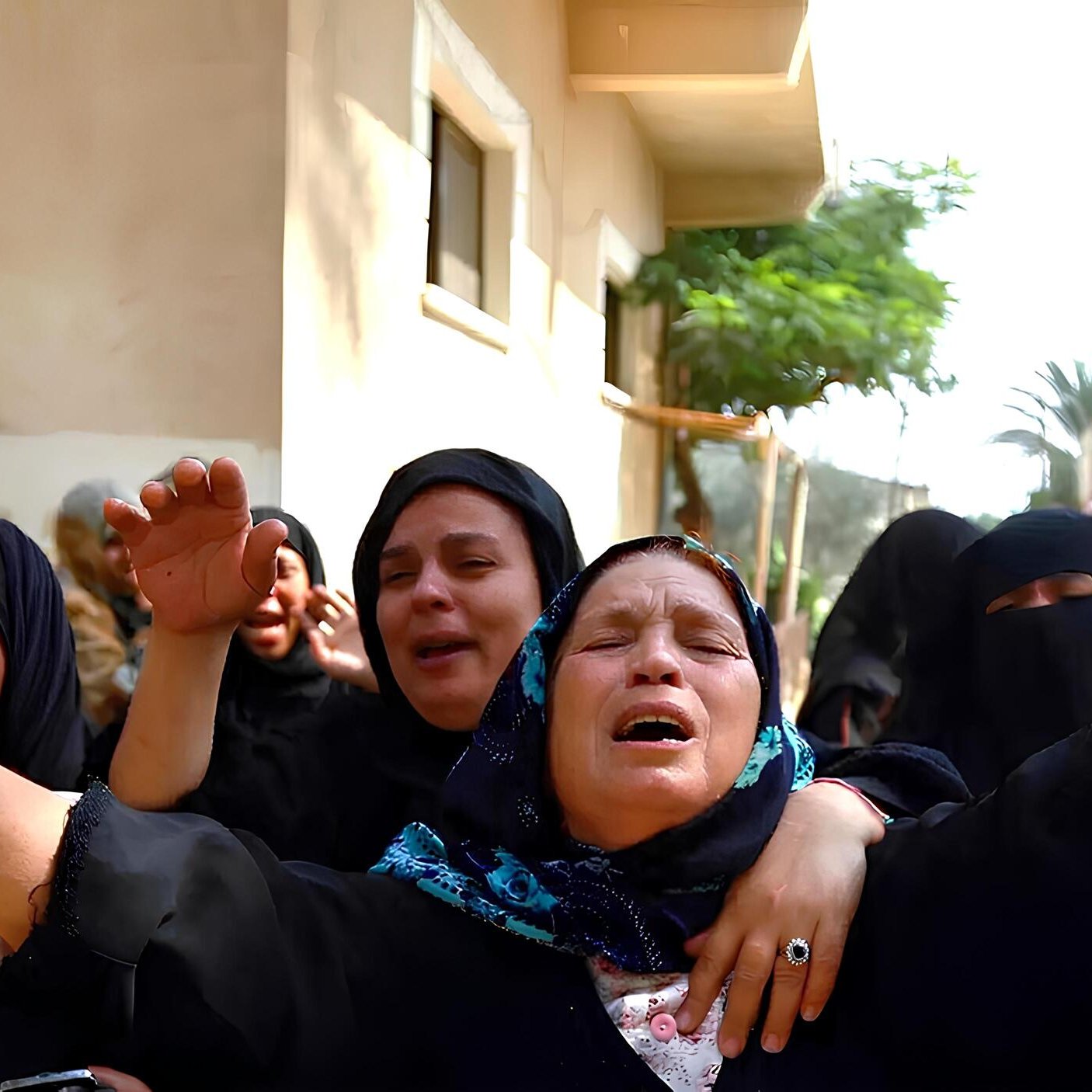 L'immagine mostra un gruppo di donne in un contesto emotivo intenso. Indossano abiti scuri e alcune di loro hanno il viso coperto. Alcune donne sembrano piangere e sollevano le braccia, mentre altre mostrano espressioni di sofferenza. In mano hanno foglie verdi, che possono rappresentare un simbolo di lutto o commemorazione. Sullo sfondo, si intravedono edifici e vegetazione. L'atmosfera è carica di emotività e partecipazione.