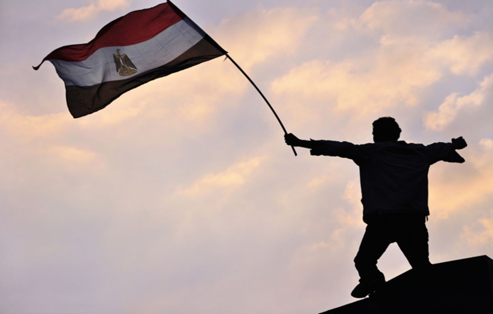 L'image montre une silhouette d'une personne debout sur une plateforme, levant un drapeau. Le drapeau est égyptien, avec des couleurs rouge, blanc, noir et le symbole du pays. L'arrière-plan présente un ciel avec des nuages aux teintes douces, créant une ambiance de détermination et de célébration. Cette scène évoque des sentiments de patriotisme et de protestation ou de victoire.