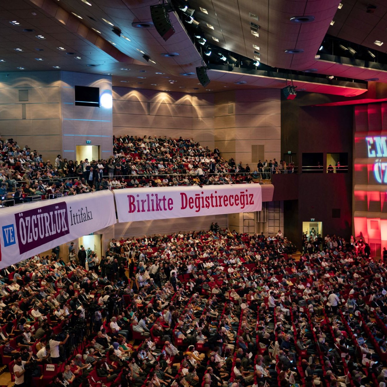 L'image montre une grande salle de conférence ou de réunion, remplie de personnes assises qui semblent assister à un événement. Les spectateurs sont disposés sur des sièges rouges, et des banderoles avec des inscriptions en turc sont accrochées sur les murs, indiquant probablement des slogans liés à des thèmes de travail ou de liberté. Un éclairage rouge illumine le fond de la scène, où des mots supplémentaires sont projetés, renforçant l'atmosphère d'un rassemblement important.