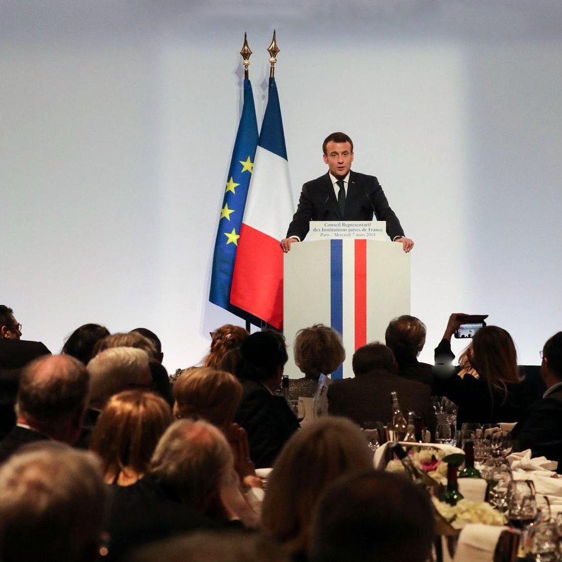 L'image montre un événement formel où une personne, probablement un homme politique, se tient devant un podium. Il parle à un public assis à des tables bien dressées, avec des couverts et des verres. En arrière-plan, on peut voir des drapeaux tricolores, bleu, blanc et rouge, représentant la France. L'atmosphère semble être celle d'un dîner officiel ou d'une réception. Les invités sont attentifs et nombreux, reflétant un moment de discours ou de présentation dans un cadre élégant.