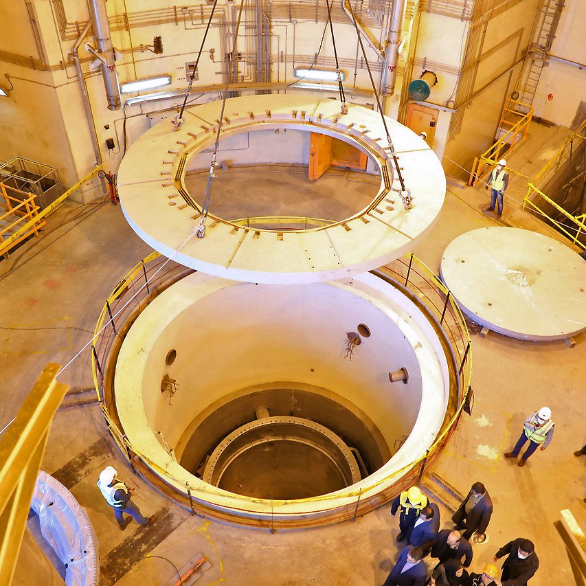 L'image montre l'intérieur d'une installation industrielle, probablement liée à la production d'énergie. On y voit un grand réacteur ou un réservoir cylindrique au centre, entouré de structures métalliques et de passerelles. Au-dessus, une grande plateforme circulaire est en train d'être soulevée, tandis qu'en bas, on aperçoit une zone vide. Plusieurs personnes en casques de sécurité se trouvent dans la pièce, probablement en train d'inspecter ou de superviser l'opération. L'éclairage est principalement artificiel, et les murs sont en béton. L'ensemble dégage une atmosphère technique et contrôlée.