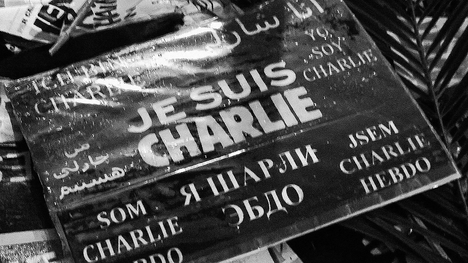 L'image montre une affiche ou un panneau avec l'inscription "JE SUIS CHARLIE" en plusieurs langues. Les mots sont écrits en noir sur fond blanc, et le tout a un aspect qui évoque le soutien et la solidarité, probablement en rapport avec les événements tragiques auxquels cette phrase fait allusion. On peut également voir des éléments environnants qui semblent être des morceaux de papier ou des affiches, ce qui donne un aspect de rassemblement ou de manifestation.