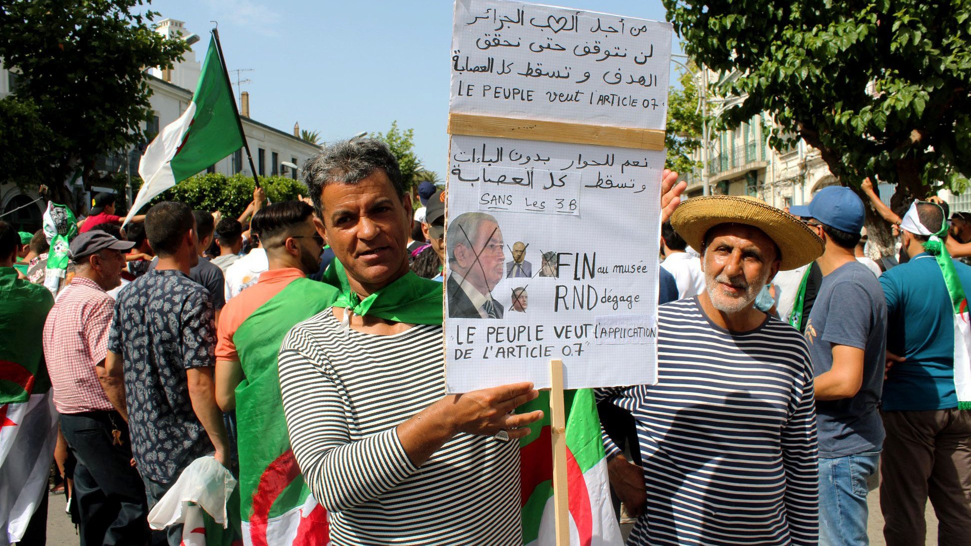 تظهر في الصورة مجموعة من الأشخاص في تجمع أو مظاهرة. يحمل بعضهم لافتات تتضمن شعارات سياسية تتعلق بالمطالب الشعبية في الجزائر. يرتدي المشاركون ملابس بألوان متنوعة، ويبدو أن هناك استخداماً للعلم الجزائري. المشهد يعكس روح الحماس والتعبير عن المطالب الجماهيرية.