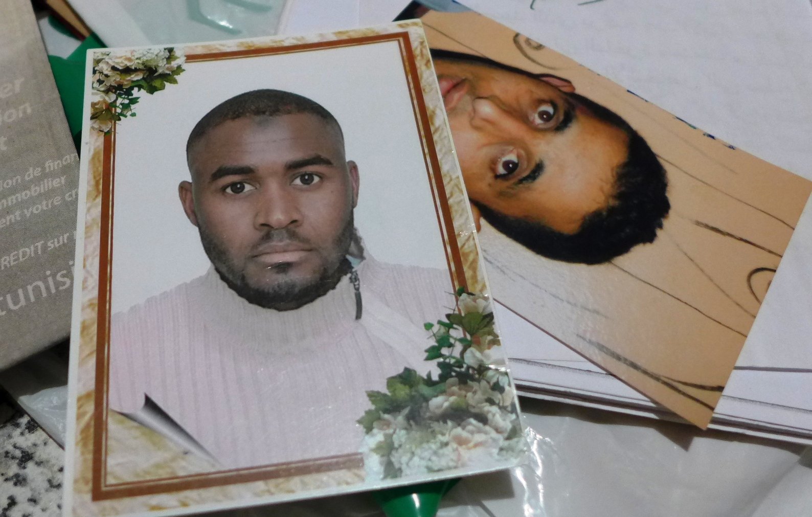 الصورة تحتوي على صورتين شخصيتين. الصورة الأولى لشخص يرتدي سترة بيضاء ويظهر تعبير وجه جدي، محاط بإطار مزخرف بالأزهار. أما الصورة الثانية، فهي لشخص آخر يظهر بشكل غير واضح، مع خلفية بسيطة. يبدو أن الصور موضوعة مع مجموعة من الأوراق أو المستندات.