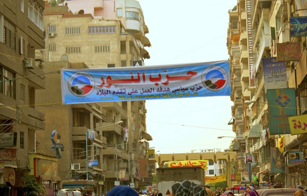 L'image montre une rue urbaine animée avec des bâtiments de plusieurs étages de chaque côté. Au-dessus de la route, il y a une grande banderole avec du texte en arabe. Cette banderole semble promouvoir un parti politique, qui pourrait être lié à des événements ou des activités locales. On aperçoit également des personnes marchant dans la rue, certaines portant des vêtements traditionnels. L'atmosphère de l'image évoque l'animation d'un marché ou d'un rassemblement communautaire.