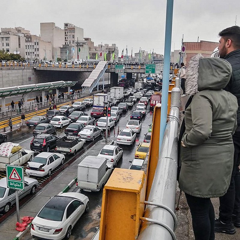 L'image montre une scène de circulation dense dans une ville. On peut voir de nombreuses voitures de différentes marques immobilisées dans un embouteillage. À l'arrière-plan, on distingue des bâtiments urbains et une route à plusieurs niveaux. Il y a des panneaux de signalisation visibles, et deux personnes se tiennent sur un balcon ou une balustrade, observant la circulation en contrebas. Le ciel est nuageux, ce qui suggère un temps gris et éventuellement pluvieux.
