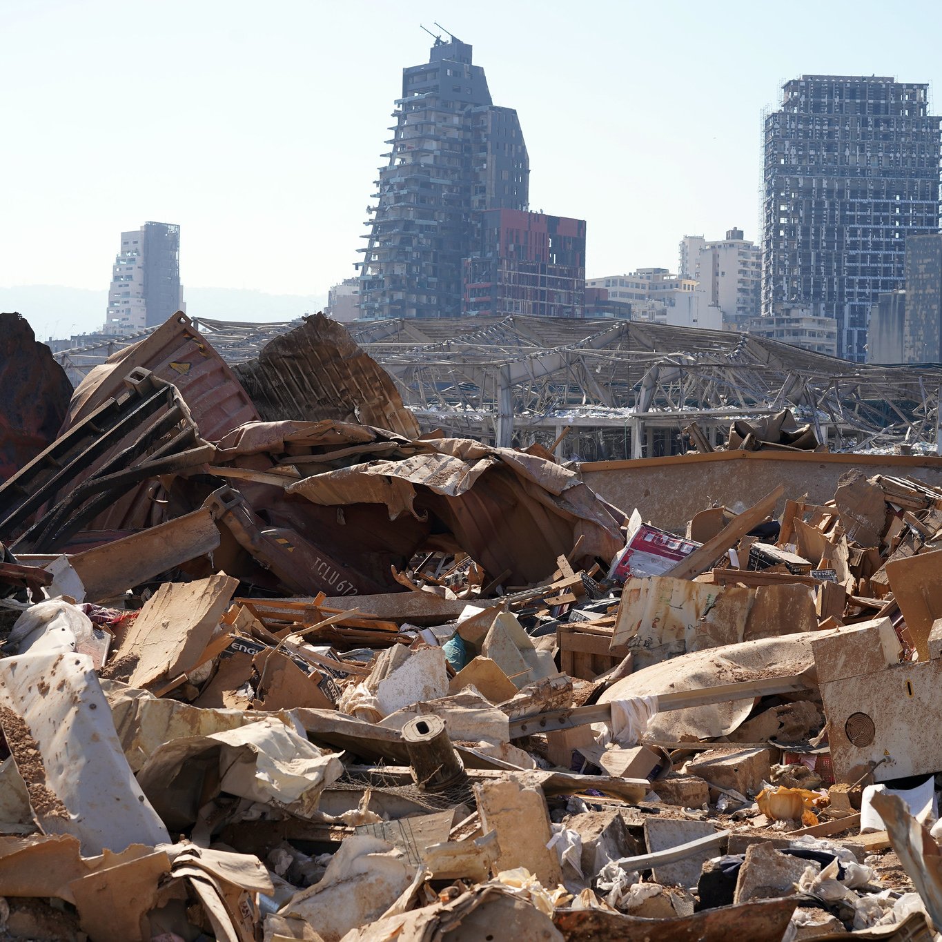 L'image montre un paysage urbain dévasté, avec des débris et des matériaux en désordre dans le premier plan. On peut voir des bâtiments partiellement détruits en arrière-plan, témoignant des dommages importants. L'atmosphère semble désolée, illustrant l'impact d'une catastrophe, probablement une explosion ou une destruction massive. Des structures modernes sont visibles, mais leur état montre les effets néfastes de l'événement. Le ciel est dégagé, contrastant avec la gravité de la scène au sol.