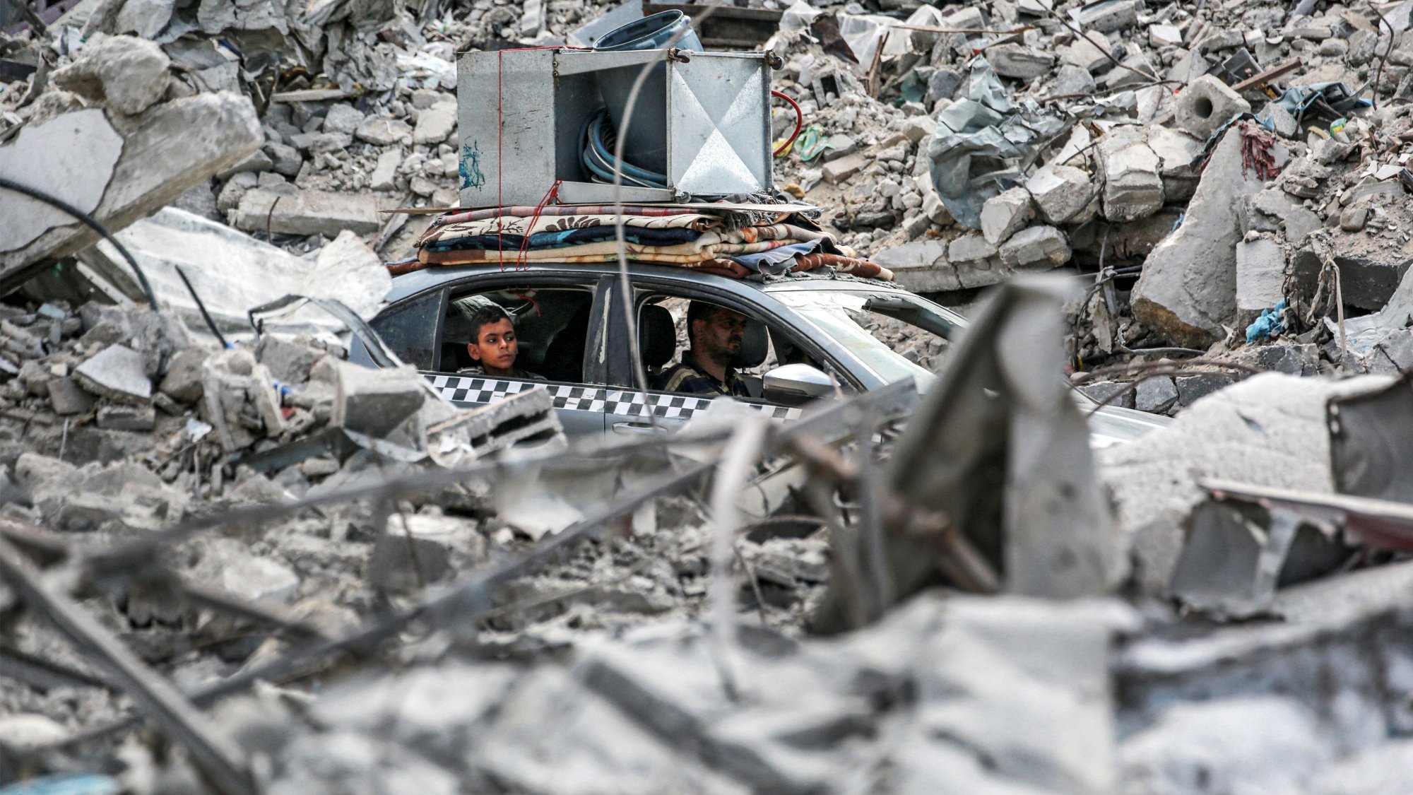 L'immagine mostra una scena di distruzione, con un'ampia quantità di macerie sparse ovunque. Al centro, c'è un'auto parcheggiata tra i detriti, con due persone sedute all'interno. Le macerie sembrano provenire da edifici crollati, evidenziando un contesto di crisi o di conflitto. L'atmosfera è carica di tensione e desolazione, con un forte contrasto tra l'umano e la devastazione circostante.