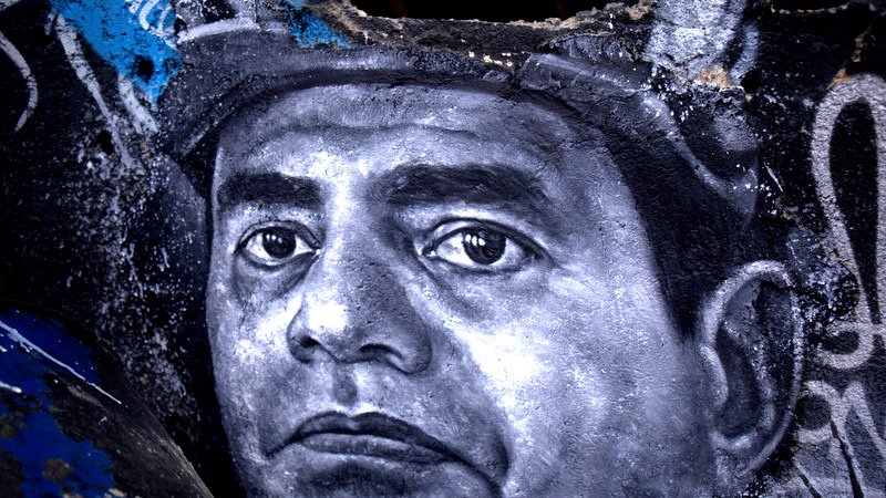 L'image représente une œuvre de street art, probablement une peinture murale, qui montre un portrait en noir et blanc d'un homme. Le visage est détaillé, avec une expression sérieuse et des yeux perçants. Le fond semble être un mur abîmé, avec des graffitis et des marques de peinture colorées, ce qui crée un contraste avec le portrait monochrome. L'ensemble donne une impression de profondeur et de caractère à l'œuvre.