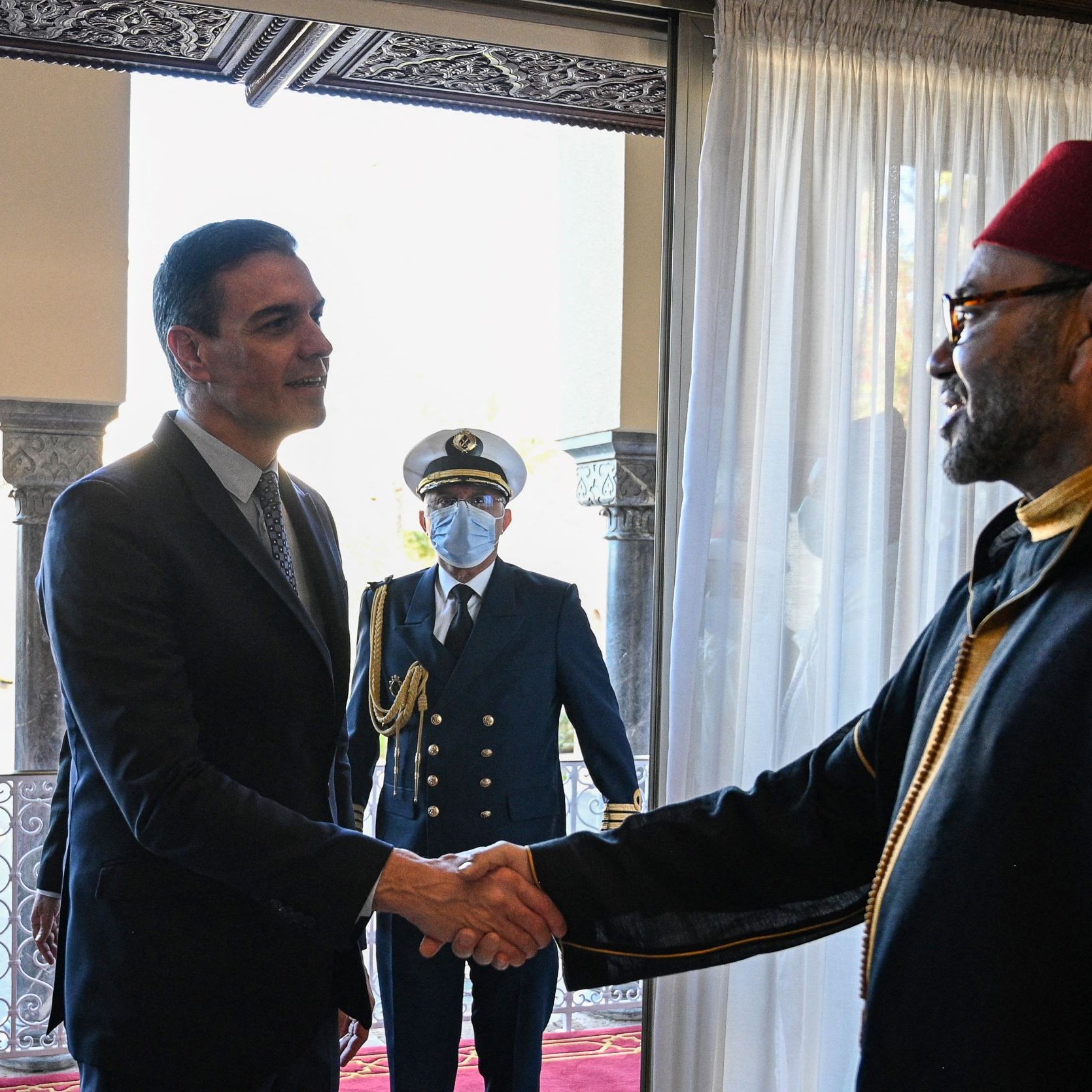 تظهر في الصورة لقاء بين شخصين، حيث يقوم أحدهما بمصافحة الآخر. يبدو أن الاجتماع رسمي، وذلك بسبب الملابس الأنيقة التي يرتديها الشخصان. في الخلفية، يوجد شخص ثالث يبدو أنه يؤدي دورًا رسميًا أيضًا. الأجواء توحي بالاحترام والتقدير بين الحضور. الأثاث وإعداد الغرفة يظهران بشكل تقليدي وفاخر.