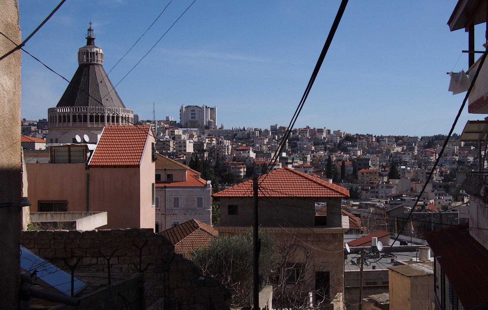 L'image montre un paysage urbain avec des bâtiments aux toits en tuiles rouges, typiques de certaines architectures méditerranéennes. On peut observer un dôme sur l'un des bâtiments, sans doute une église ou un édifice religieux. En arrière-plan, la ville s'étend sur plusieurs niveaux, avec de nombreuses constructions. Le ciel est dégagé et ensoleillé, créant une ambiance lumineuse. Des lignes électriques traversent l'image, ajoutant un élément urbain au paysage.