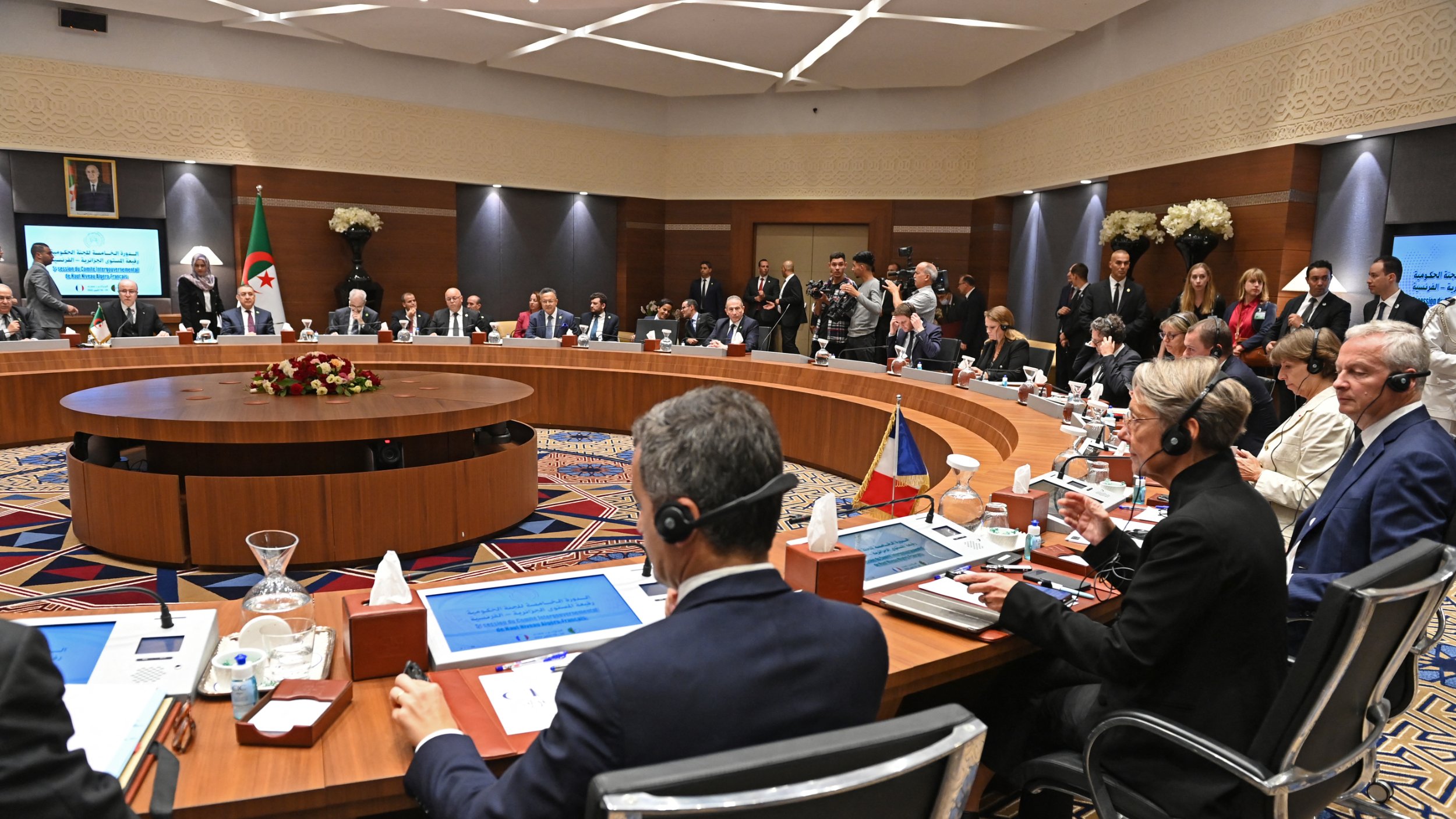 تظهر الصورة اجتماعًا يُعقَد في قاعة مؤتمرات، حيث يجلس العديد من المشاركين حول طاولة دائرية. تتواجد شاشات عرض أمام بعض الأشخاص، وتبدو القاعة مزينة ببعض الزهور. يُلاحظ وجود عدد من الحضور في الخلفية، مما يشير إلى أهمية الحدث. المشاركون يرتدون ملابس رسمية ويظهرون في لحظة من النقاش أو الاستماع.