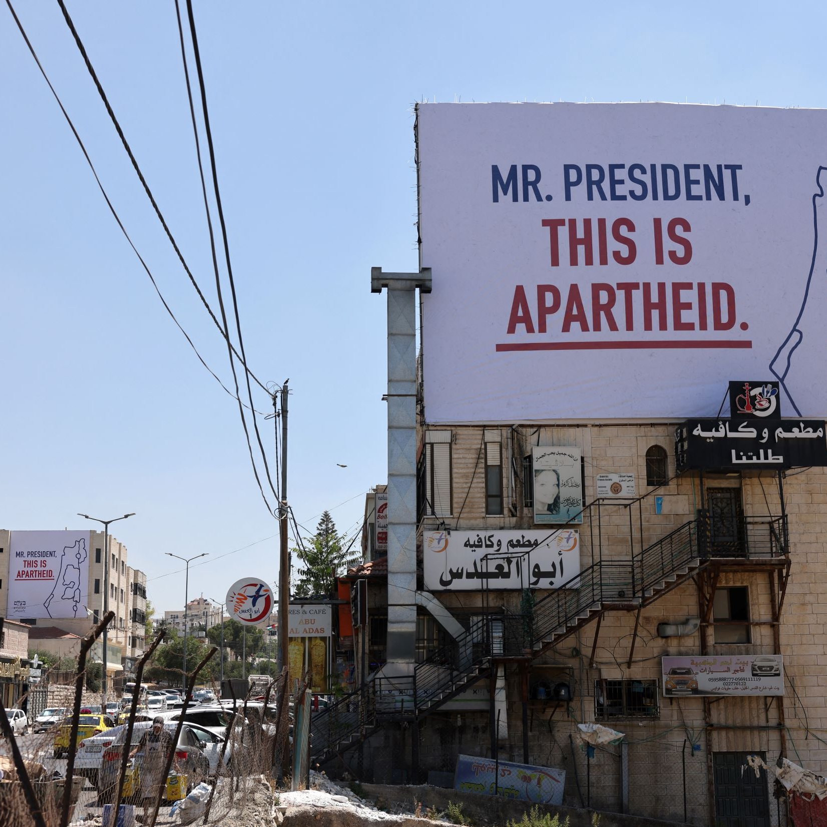 L'image montre un panneau publicitaire accroché sur un bâtiment, avec un message qui dit "MR. PRESIDENT, THIS IS APARTHEID." On peut voir des infrastructures urbaines environnantes, telles que des routes, des bâtiments et des fils de téléphone. Le contexte suggère une critique sociale ou politique liée à la situation en Israël-Palestine. Le paysage est ensoleillé et présente une ambiance de ville.