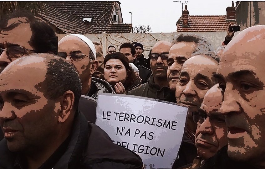 L'image montre un groupe de personnes rassemblées lors d'une manifestation. Ils semblent préoccupés et écoutent attentivement, avec des expressions sérieuses. Certains tiennent des pancartes, dont une qui porte le message "LE TERRORISME N'A PAS DE RELIGION". L'environnement semble urbain, avec des bâtiments en arrière-plan. L'atmosphère suggère une revendication ou une protestation sur un sujet important.