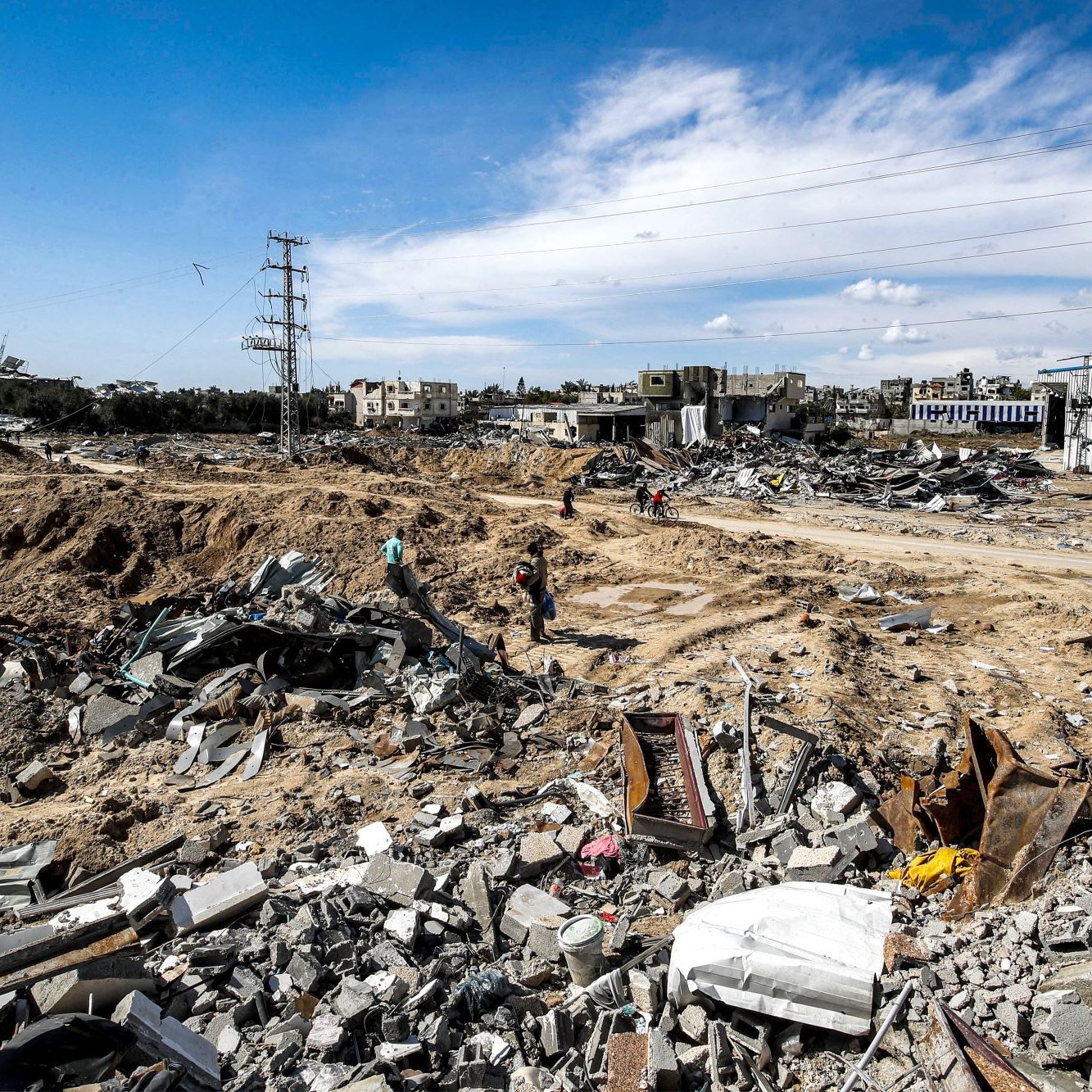 L'immagine mostra un paesaggio devastato, caratterizzato da macerie e detriti. Il terreno sembra essere stato scavato e distrutto, con edifici in rovina visibili sullo sfondo. I resti di strutture crollate sono sparsi ovunque, mentre alcune persone si muovono tra le macerie. Il cielo è parzialmente nuvoloso, e ci sono segnali di infrastrutture elettriche nei dintorni. L'atmosfera trasmette un senso di desolazione e distruzione.
