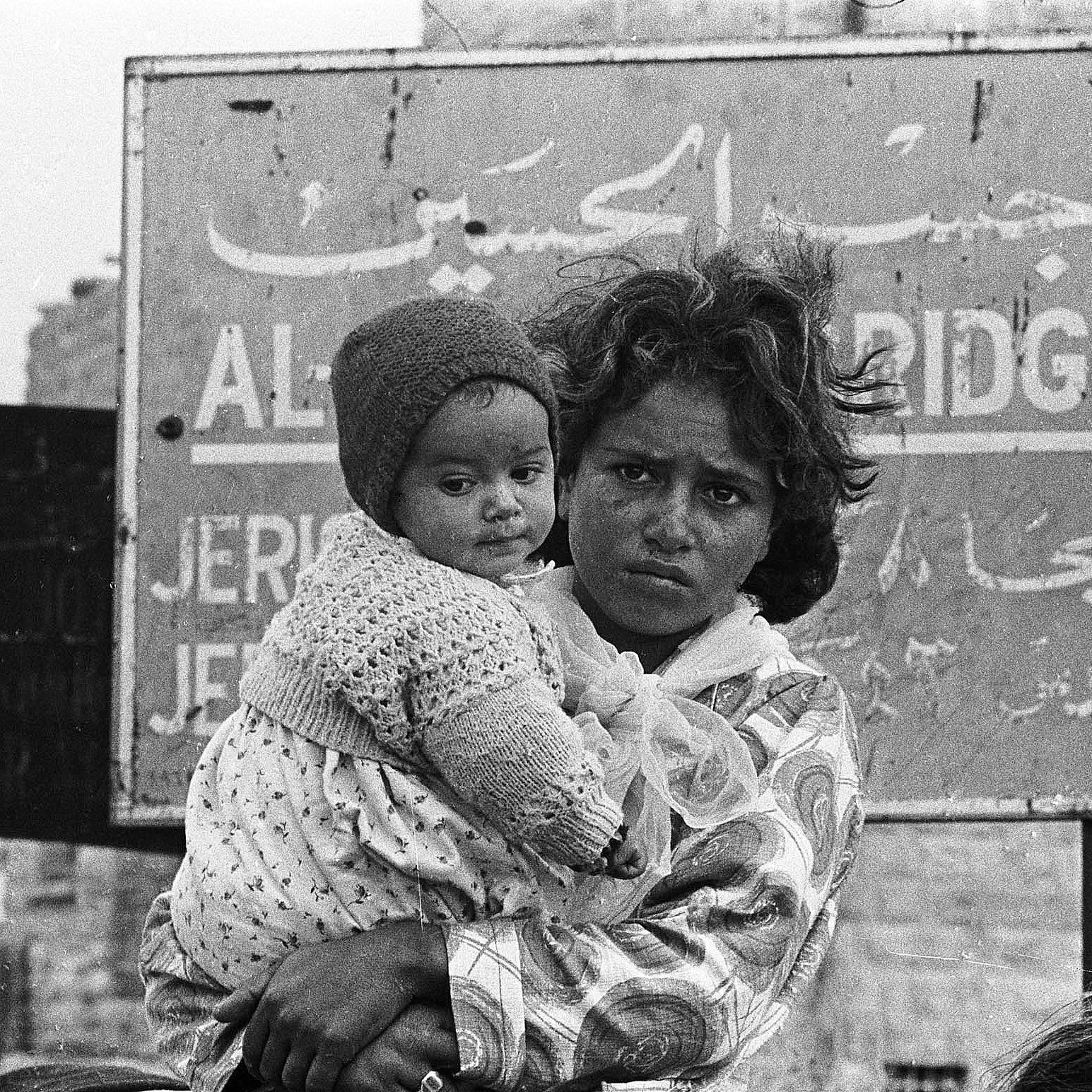 L'image montre une jeune mère, tenant dans ses bras un bébé qui porte un bonnet. La mère a un regard sérieux et semble préoccupée. En arrière-plan, on aperçoit un panneau avec des inscriptions en arabe et en anglais, indiquant des informations sur un lieu. L'atmosphère est intense, reflétant une situation de vulnérabilité. Le noir et blanc de l'image accentue l'émotion et le contexte difficile dans lequel ces personnes se trouvent.