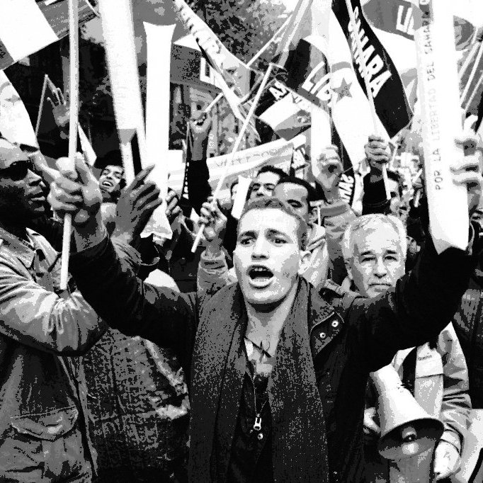 L'image montre une foule en pleine manifestation. On peut voir des personnes brandissant des drapeaux et des pancartes, exprimant leurs opinions avec passion. Au premier plan, un jeune homme s'écrie, tandis que d'autres personnes autour de lui semblent également mobilisées et engagées. L'ambiance générale dégage une forte énergie et un sentiment de solidarité parmi les manifestants. Les visages reflètent une gamme d'émotions, allant de la détermination à l'enthousiasme.